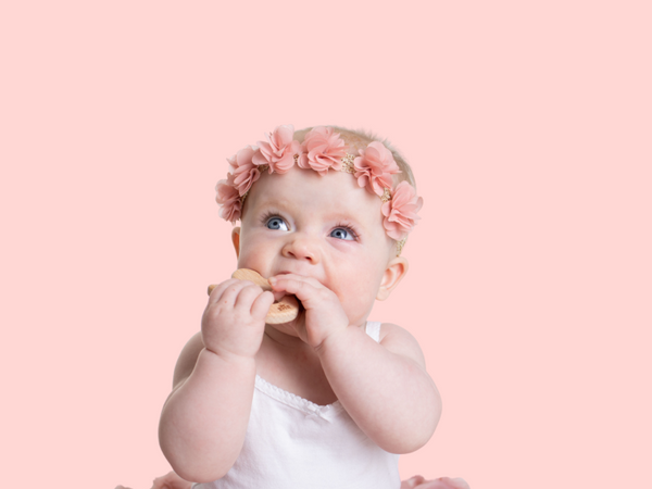 cute baby wearing a flower crown while chewing on a wooden teether in the shape of an angel.