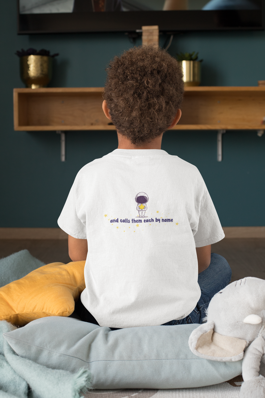 Young boy sitting on cushion with back of white kids Christian tshirt that says "and calls them each by name" below an astronaut holding a star.